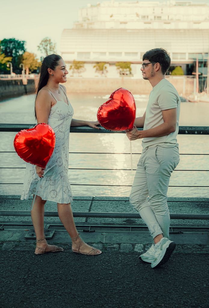 A couple with heart-shaped balloons enjoy a romantic moment by a riverside railing.