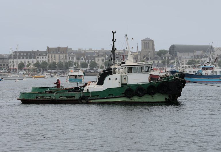 A small fishing boat moored at a busy harbor with cityscape in the background.
